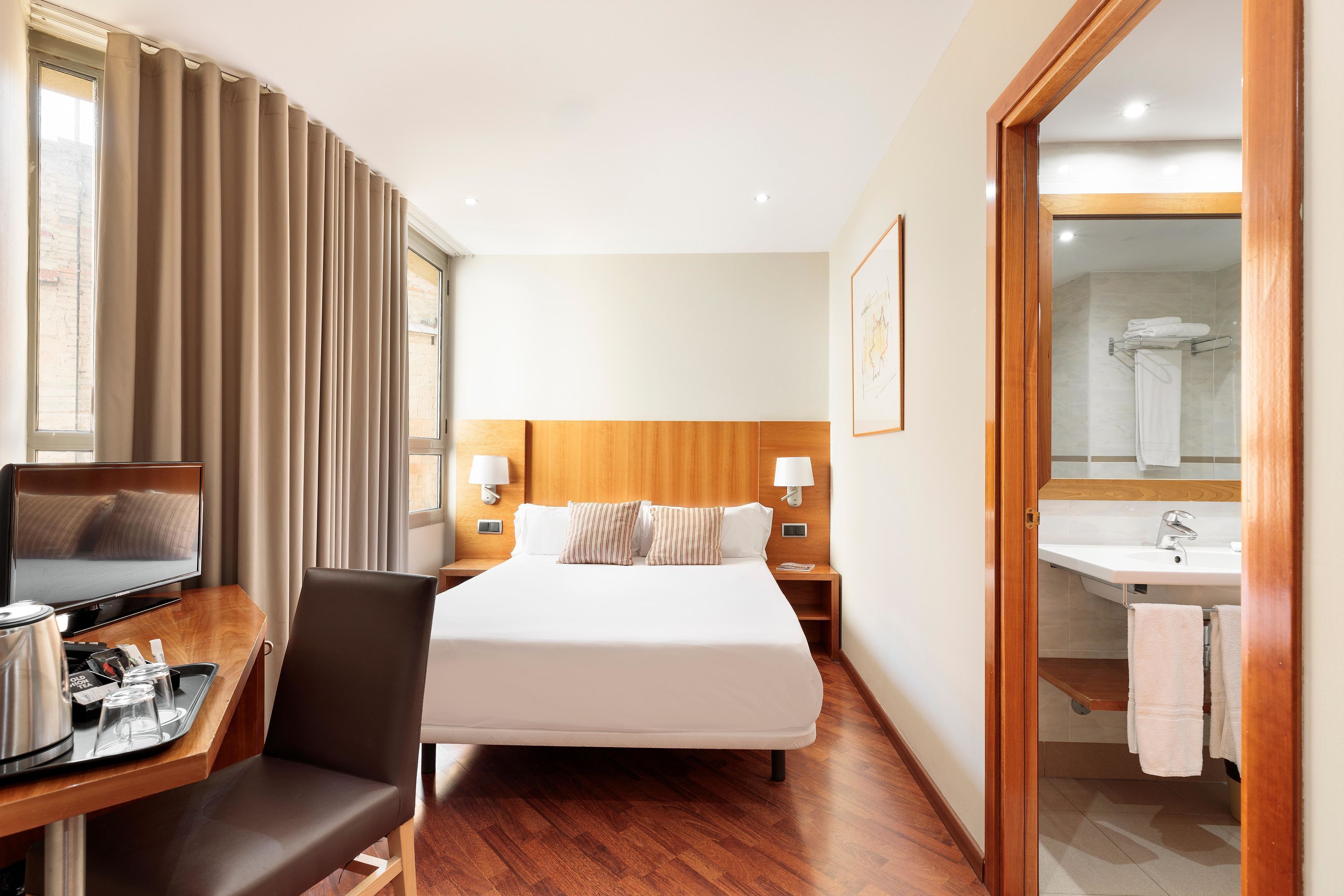Ein Hotelzimmer mit Bett, Schreibtisch mit Fernseher, Stuhl, Holz-Kopfteil, Nachttischlampen und einem durchgegangene Tür sichtbaren Badezimmer.