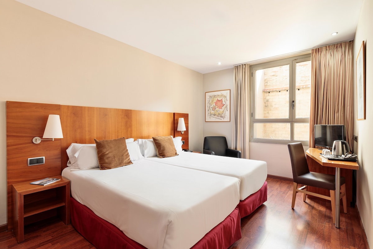 A hotel room with two beds, a desk, a chair, a TV, and a window with curtains.