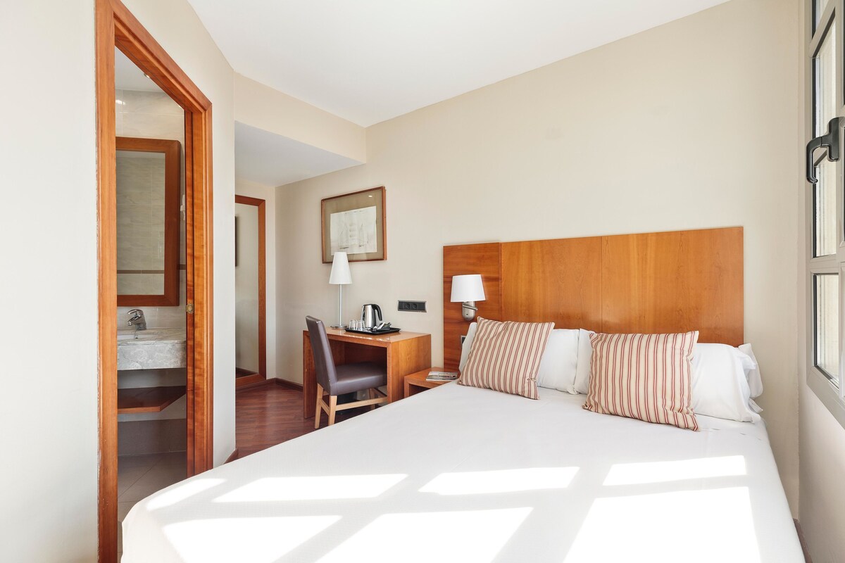 Habitación de hotel con una cama grande, cabecera de madera, escritorio con silla, mesita de noche y una ventana.
