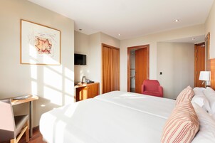 Quarto de hotel com uma cama grande, uma escrivaninha com cadeira, uma televisão e um guarda-roupa de madeira.