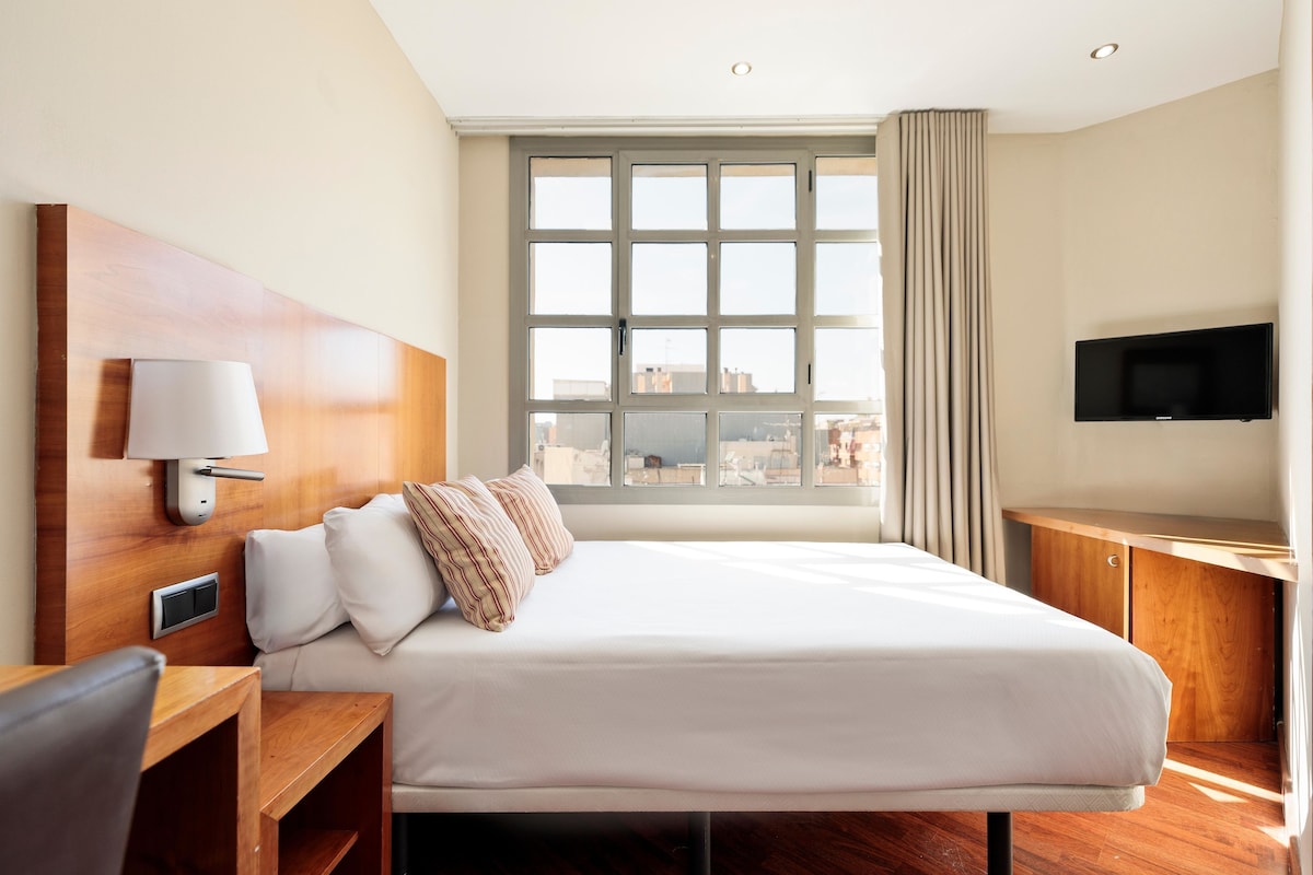 A hotel room with a large bed, a desk, a chair, a TV, and a window with a city view.