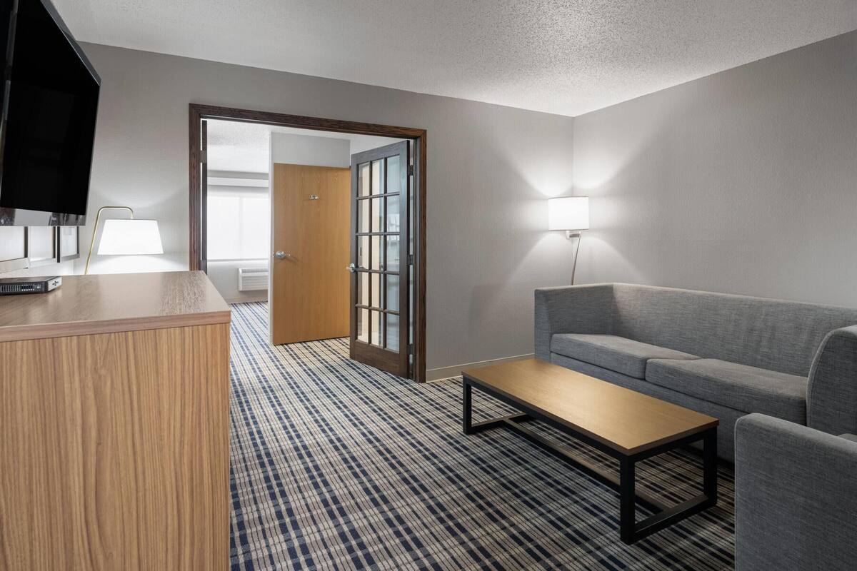 A hotel room with a wooden desk, a grey sofa, a small table, a TV, and a lamp.