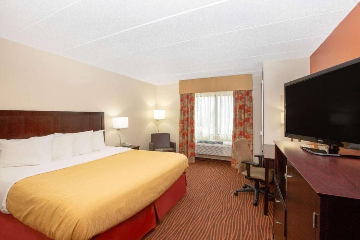 A hotel room with a large bed, a desk with a flat-screen TV, a chair, and a lamp.