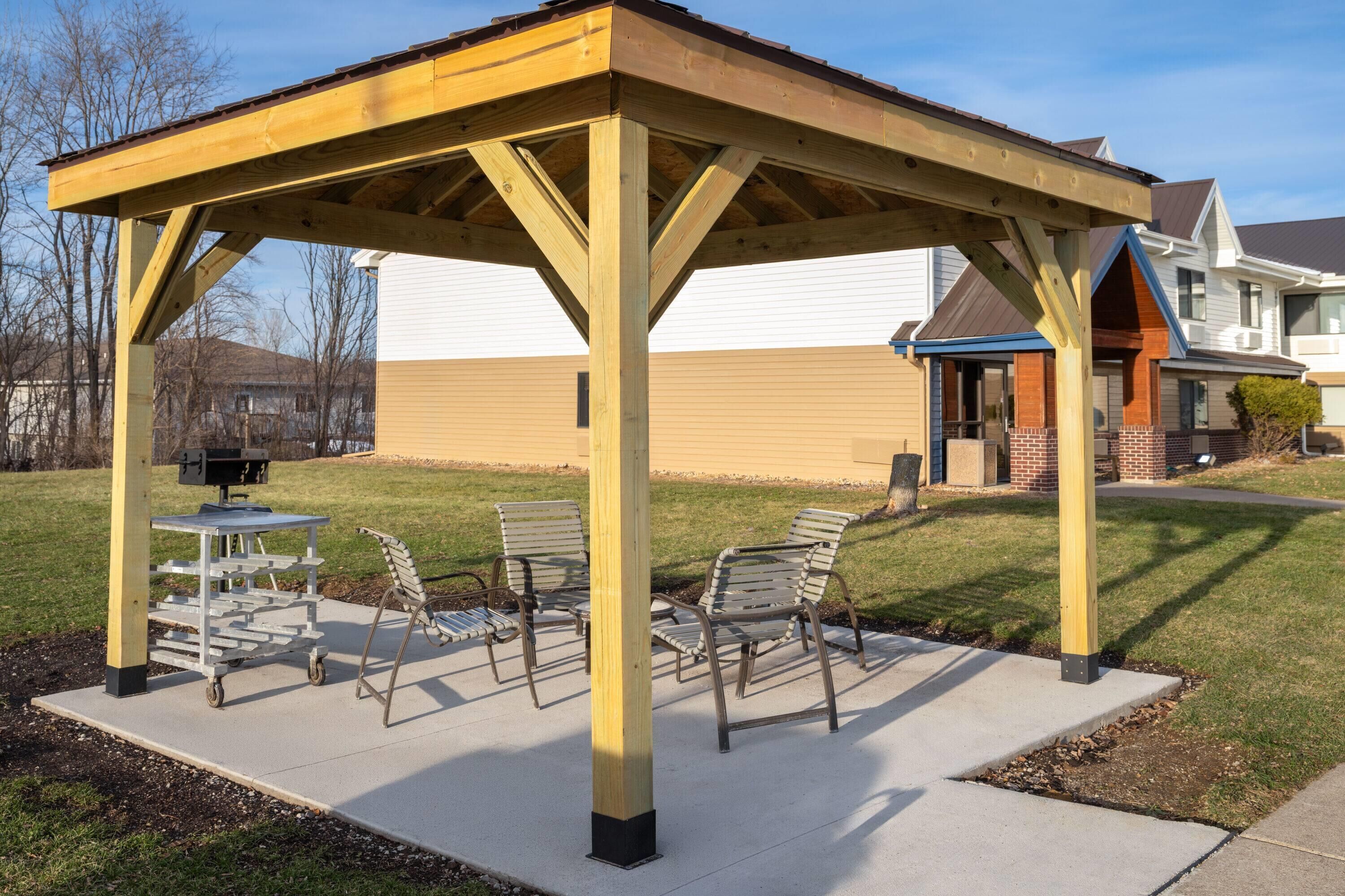 A covered outdoor seating area with a table and chairs, a grill, and a building in the background.