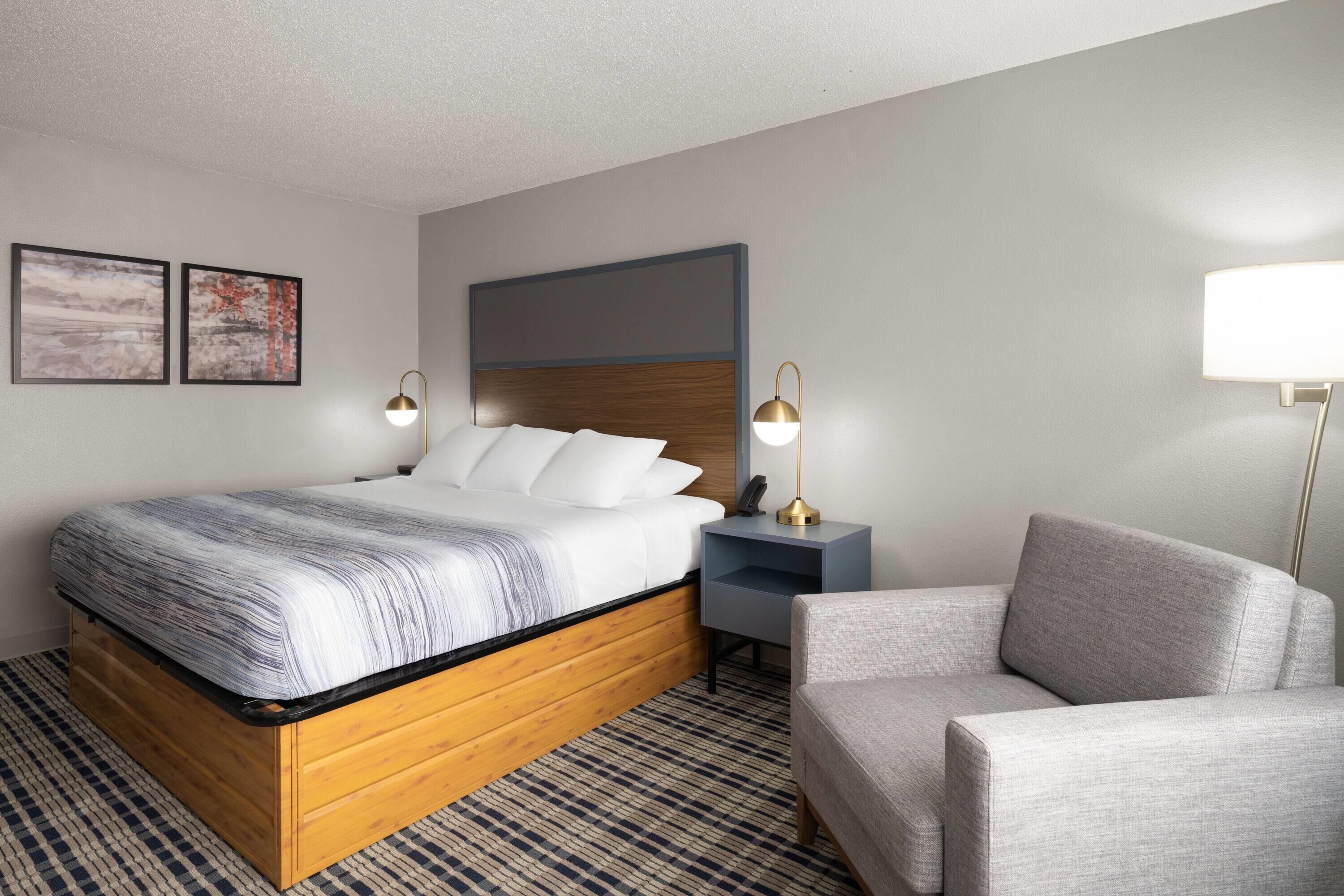 Ein Hotelzimmer mit einem hölzernen Bett, einem grauen Sessel, einem Nachttisch mit Lampe und zwei gerahmten Bildern an der Wand.