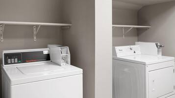 Une buanderie équipée de deux machines à laver et deux sèche-linge industriels blancs, un panneau de commande situé à gauche et une étagère au-dessus.