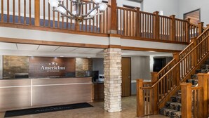 A reception area with a wooden reception desk, a stone pillar, and a staircase leading to an upper level. - AmericInn by Wyndham Muscatine (Muscatine)