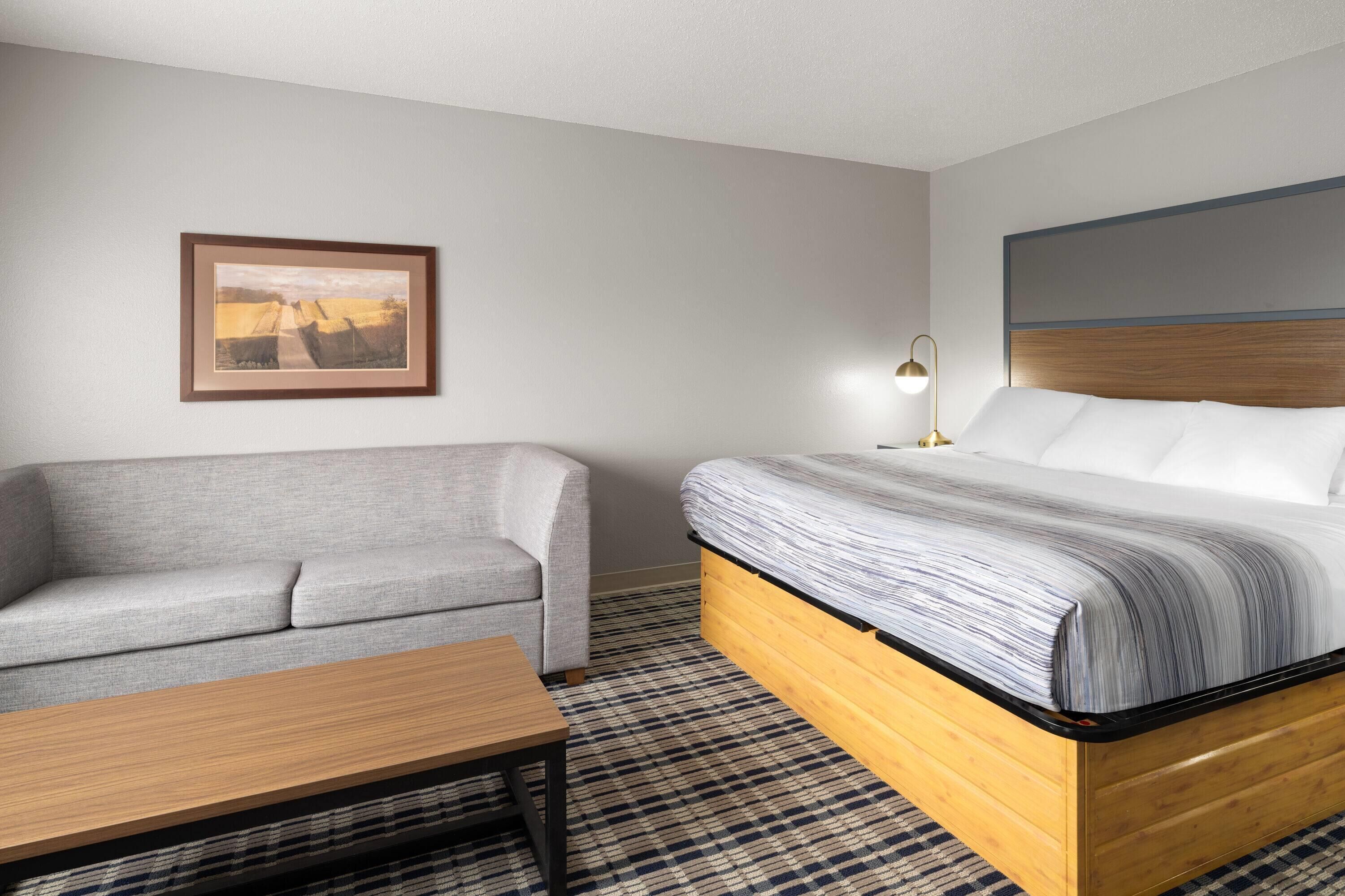 A hotel room with a bed, a gray sofa, a wooden table, and a framed picture on the wall.