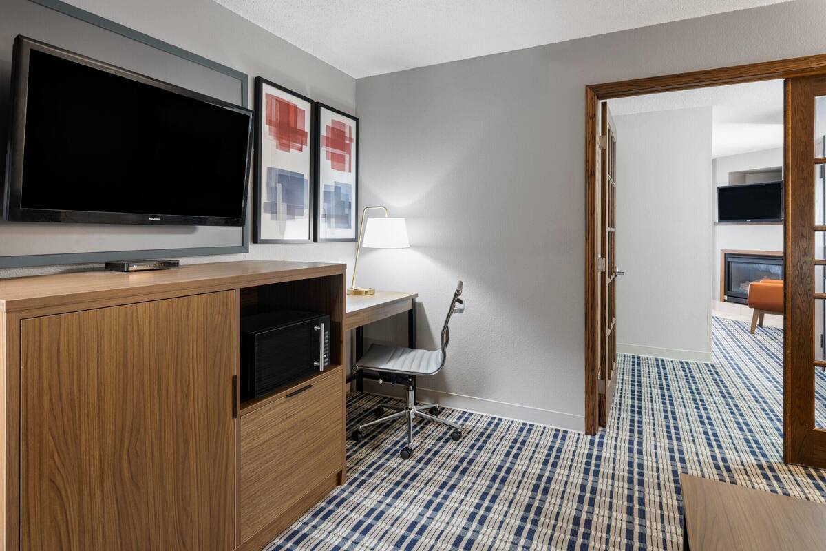 A hotel room with a large flat-screen TV, a desk with a lamp, a chair, and a patterned carpet.