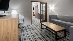 A modern hotel room with a flat-screen TV, a wooden desk, a grey sofa, a patterned carpet, and a wooden-framed door leading to another room. - AmericInn by Wyndham Muscatine (Muscatine)