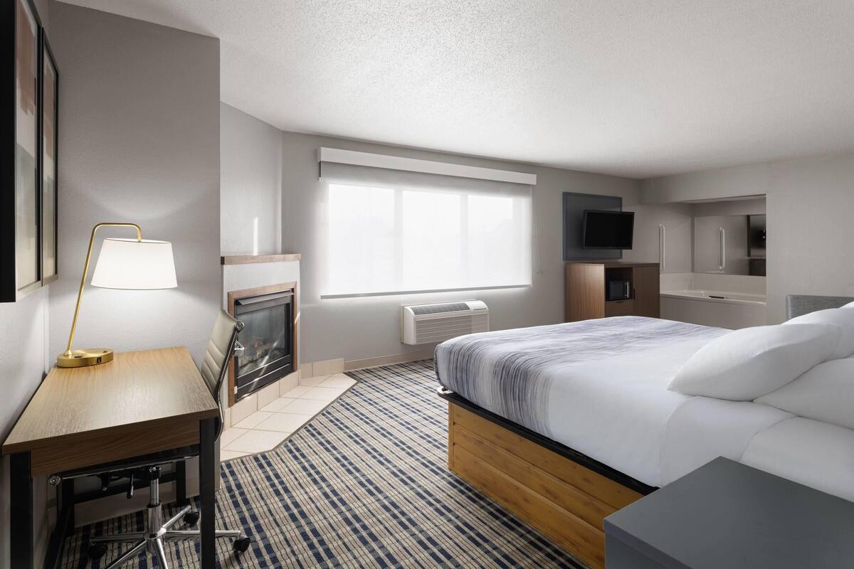 A modern hotel room with a bed, a desk, a lamp, a fireplace, a television, and a view of the city.