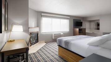 A modern hotel room with a bed, a desk, a lamp, a fireplace, a television, and a view of the city.