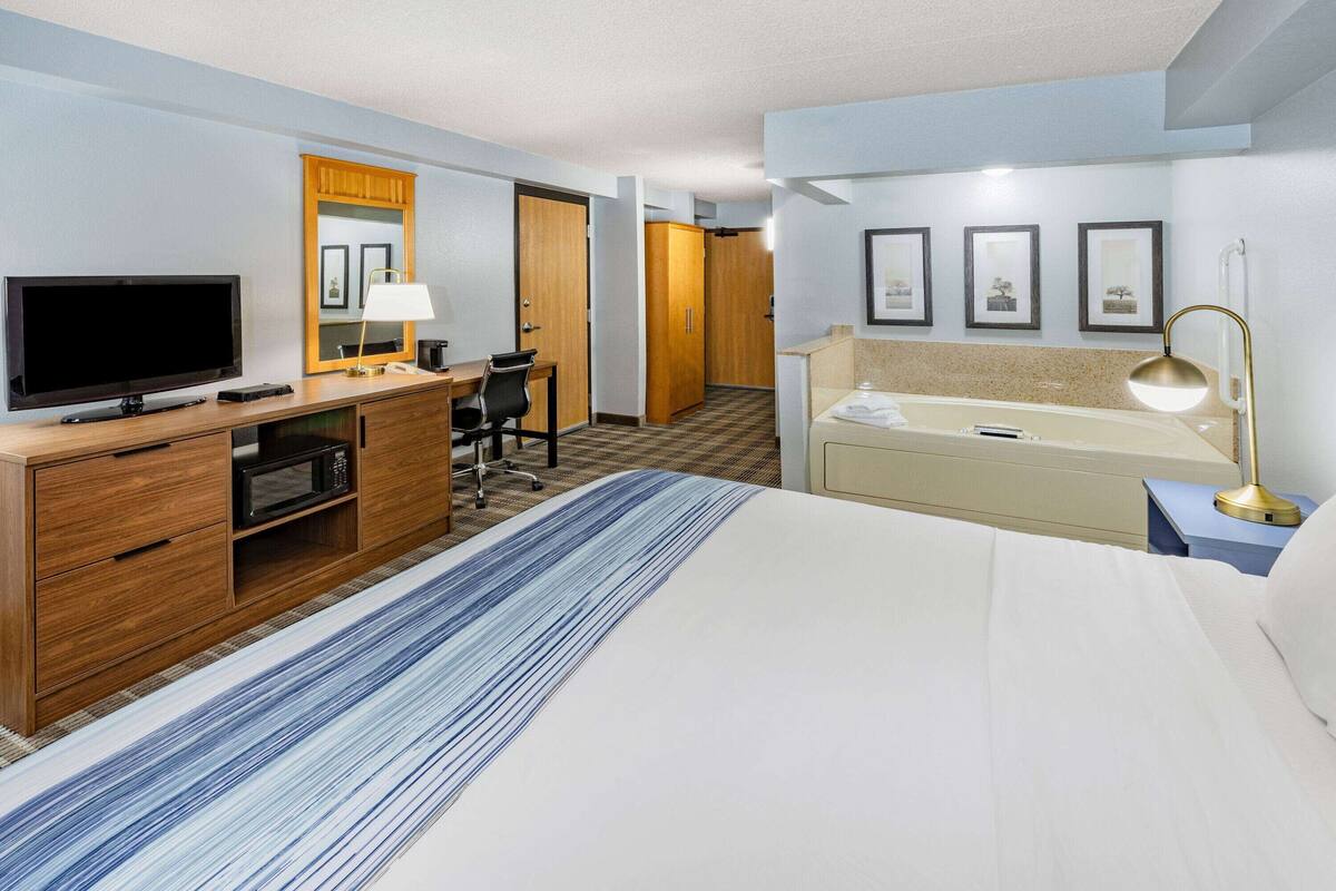 A hotel room with a bed, a desk with a TV, a microwave, a chair, a desk lamp, a vanity with a mirror, and framed pictures on the wall.
