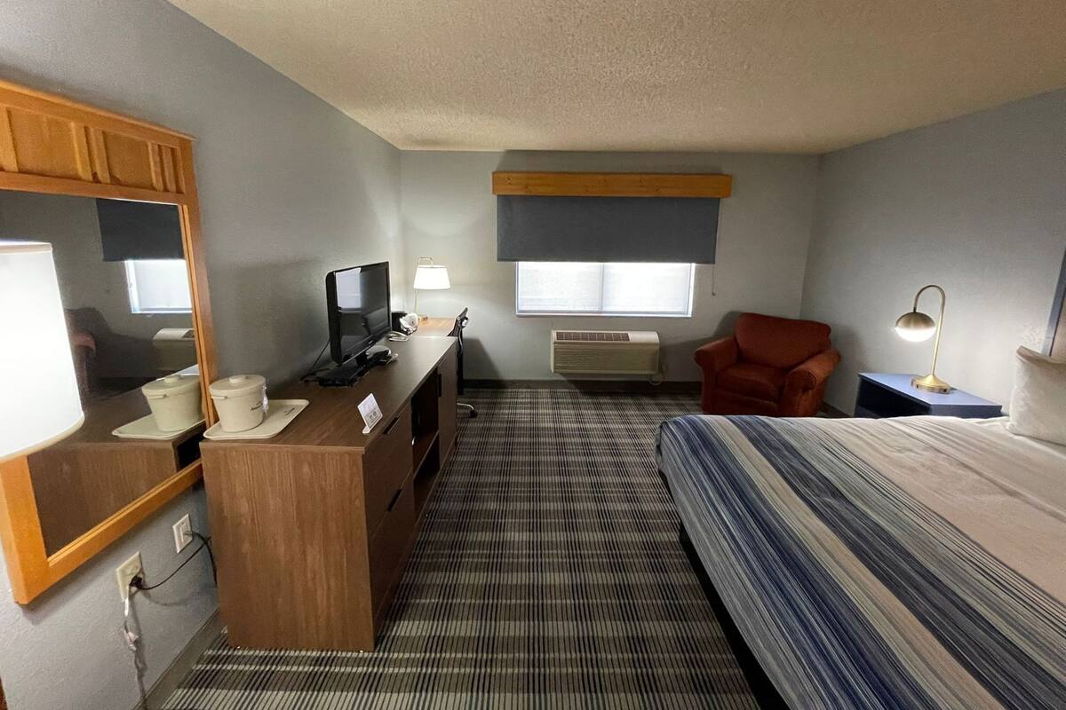 A hotel room with a bed, a desk with a computer, a chair, a mirror, a window, and an air conditioning unit.