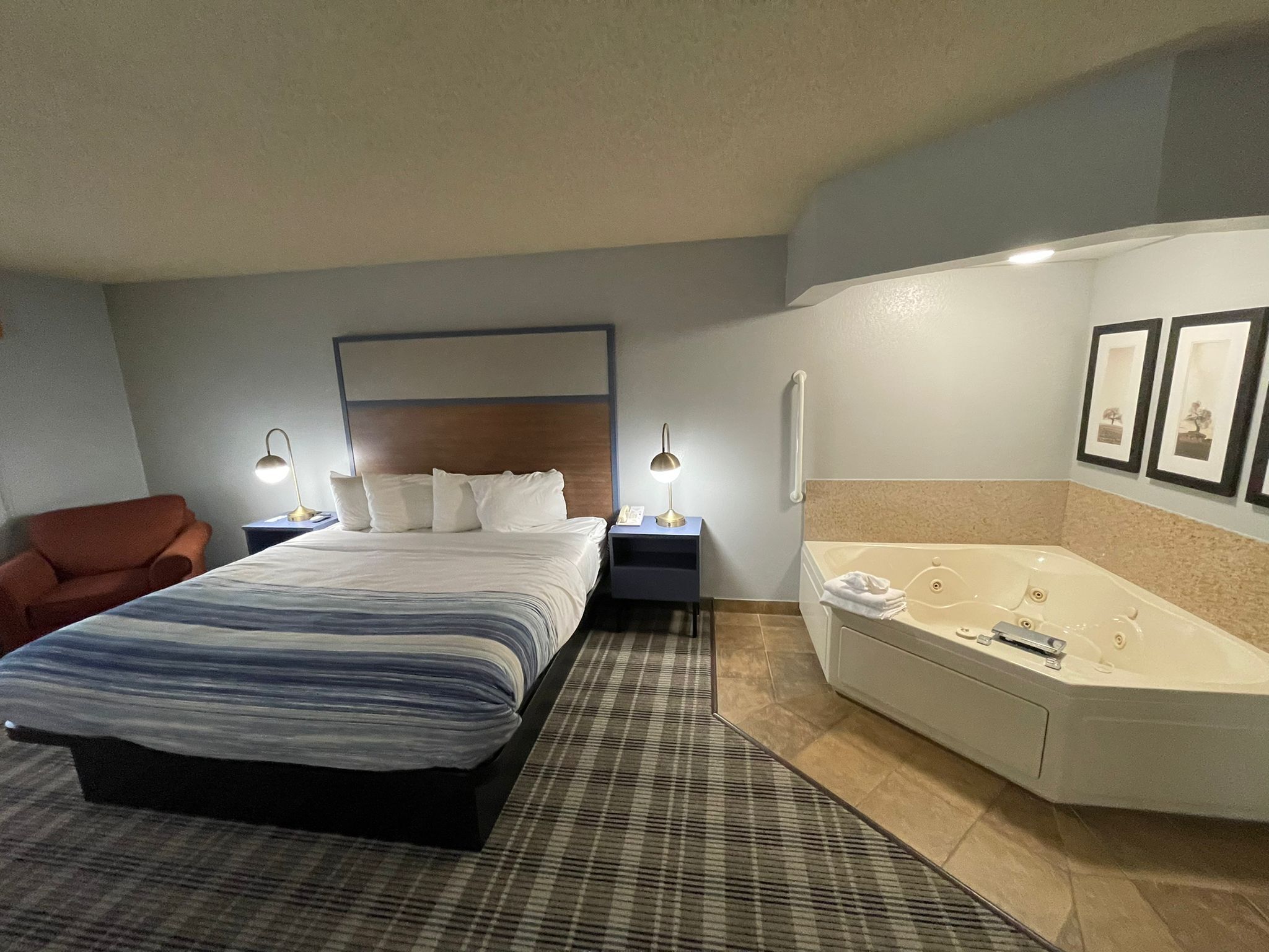 Habitación de hotel con cama, una silla, mesita de noche con lámpara, baño con bañera y cuadros en la pared.