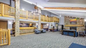 A lobby with a stone pillar, wooden railings, a reception desk, and a fireplace. - AmericInn by Wyndham Sayre (Sayre)