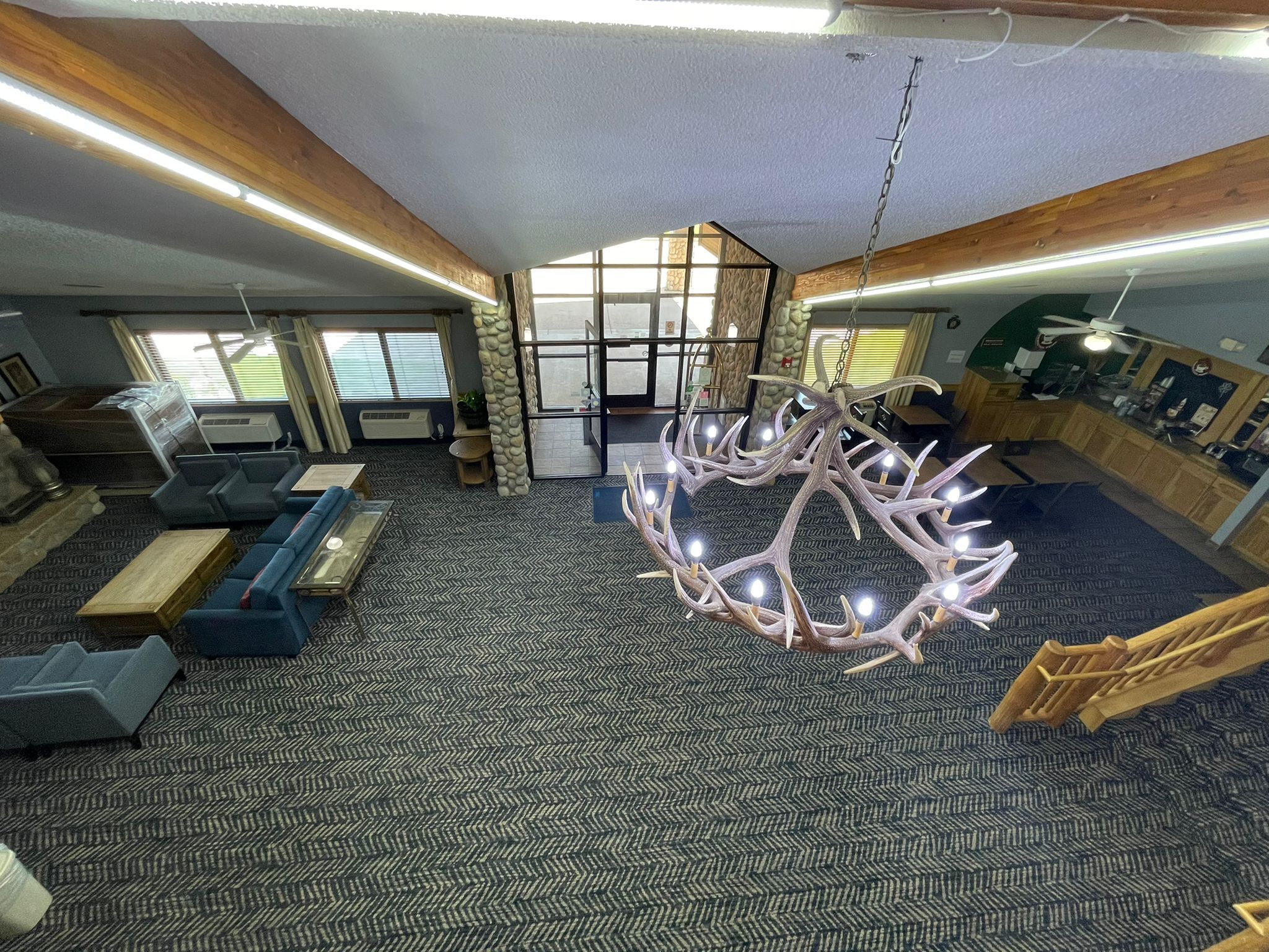 A spacious room with a herringbone carpet, a chandelier with antler-like light fixtures, and a view of the outdoors through a large window.