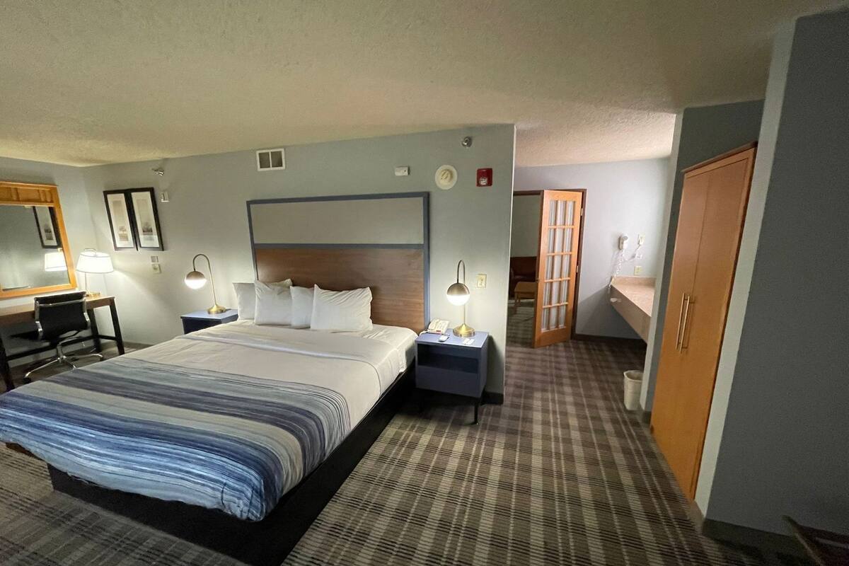 Een hotelkamer met een groot bed, nachtkastjes met lampen, een bureau, een stoel, een kledingkast en een deur die naar een andere kamer leidt.