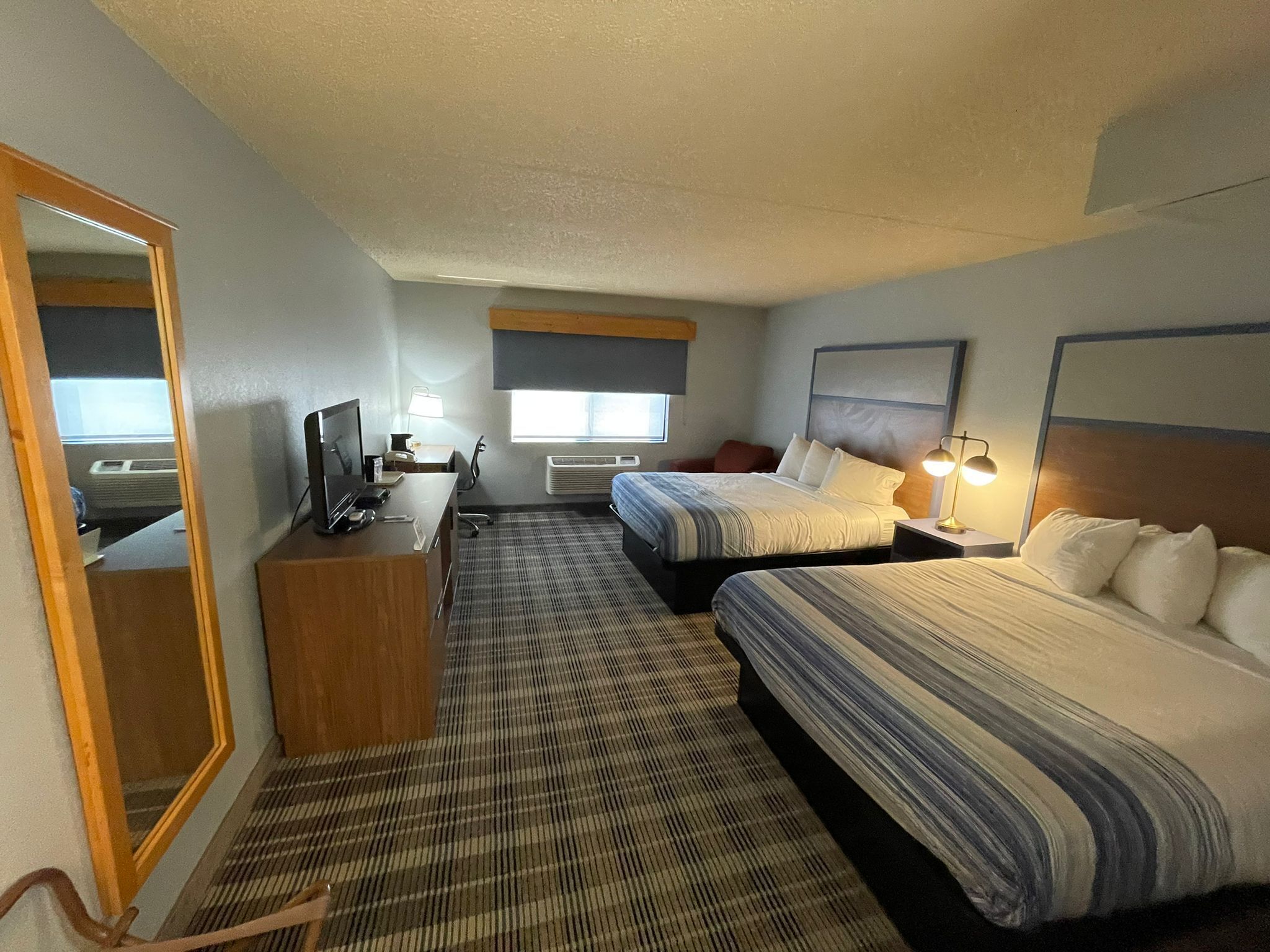 A hotel room with two beds, a desk with a computer, a mirror, and a window with blinds.