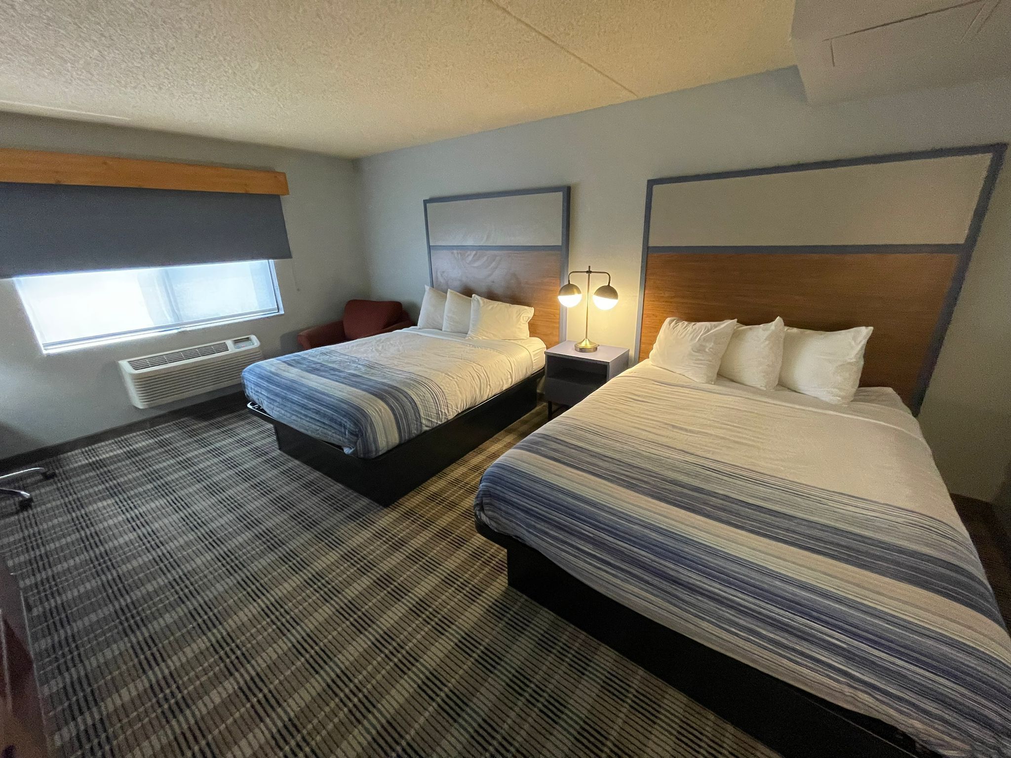 A hotel room with two beds, a window with blinds, a desk with a lamp, and a radiator.