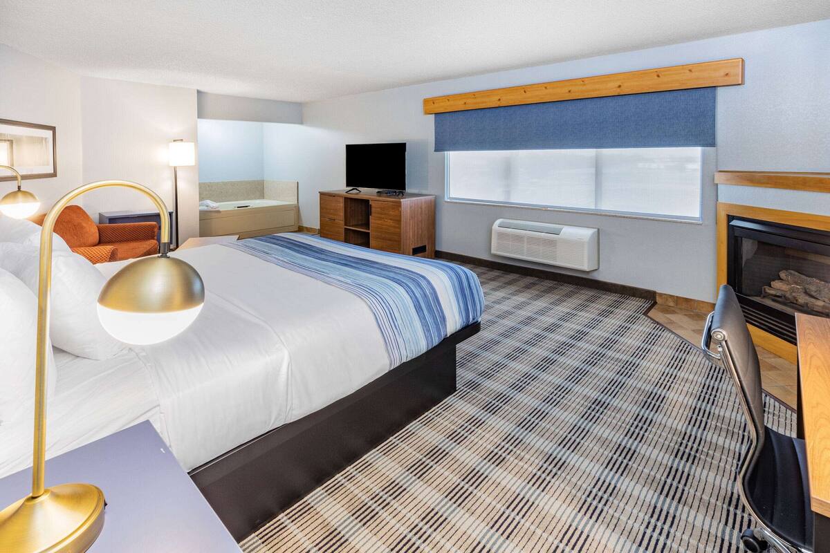 A hotel room with a bed, a desk, a chair, a television, a fireplace, and a window with blinds.