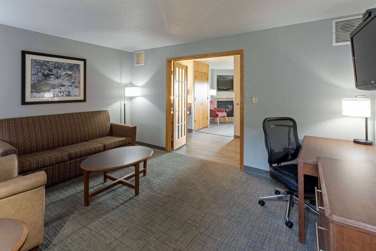 A hotel room with a sofa, a small table, a desk with a chair, a TV, and a fireplace.