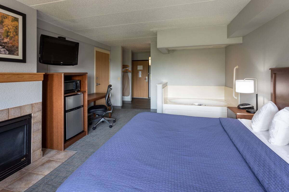 A hotel room with a large bed, a flat-screen TV mounted above the fireplace, a desk with a computer, and a shower area.