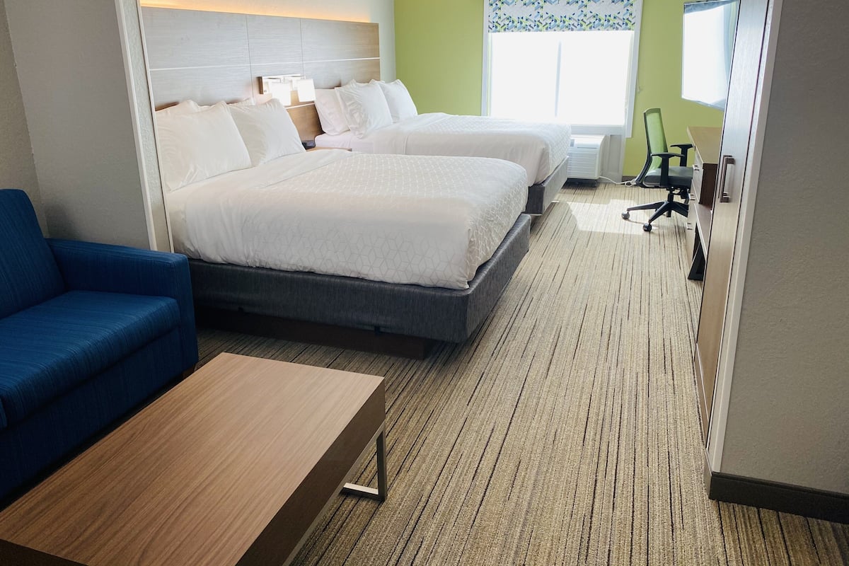 Habitación de hotel con dos camas, un sofá azul, una mesa de centro de madera, un escritorio y una ventana con cortinas estampadas.