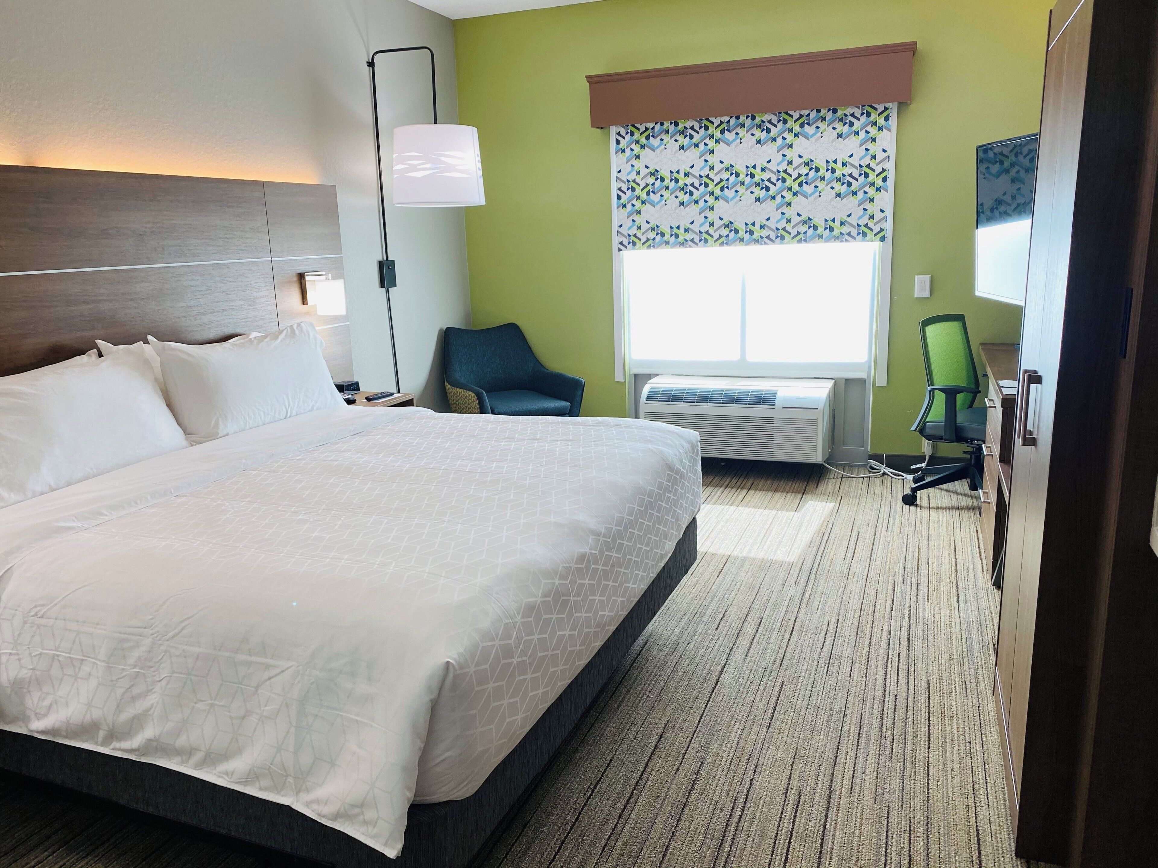 A hotel room with a large bed, a desk, a chair, a window with blinds, and an air conditioning unit.
