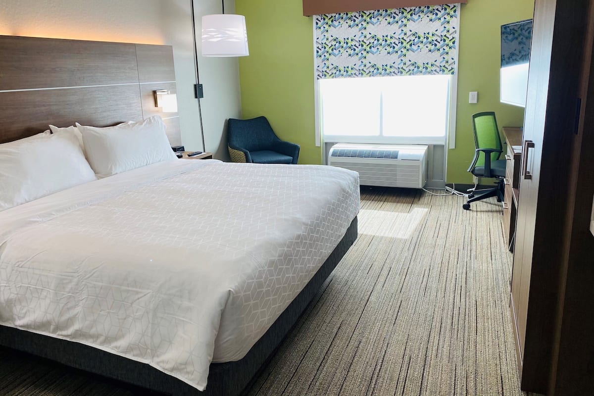 Habitación de hotel con una cama grande, un escritorio con silla, una lámpara de pie y una ventana con cortinas estampadas.