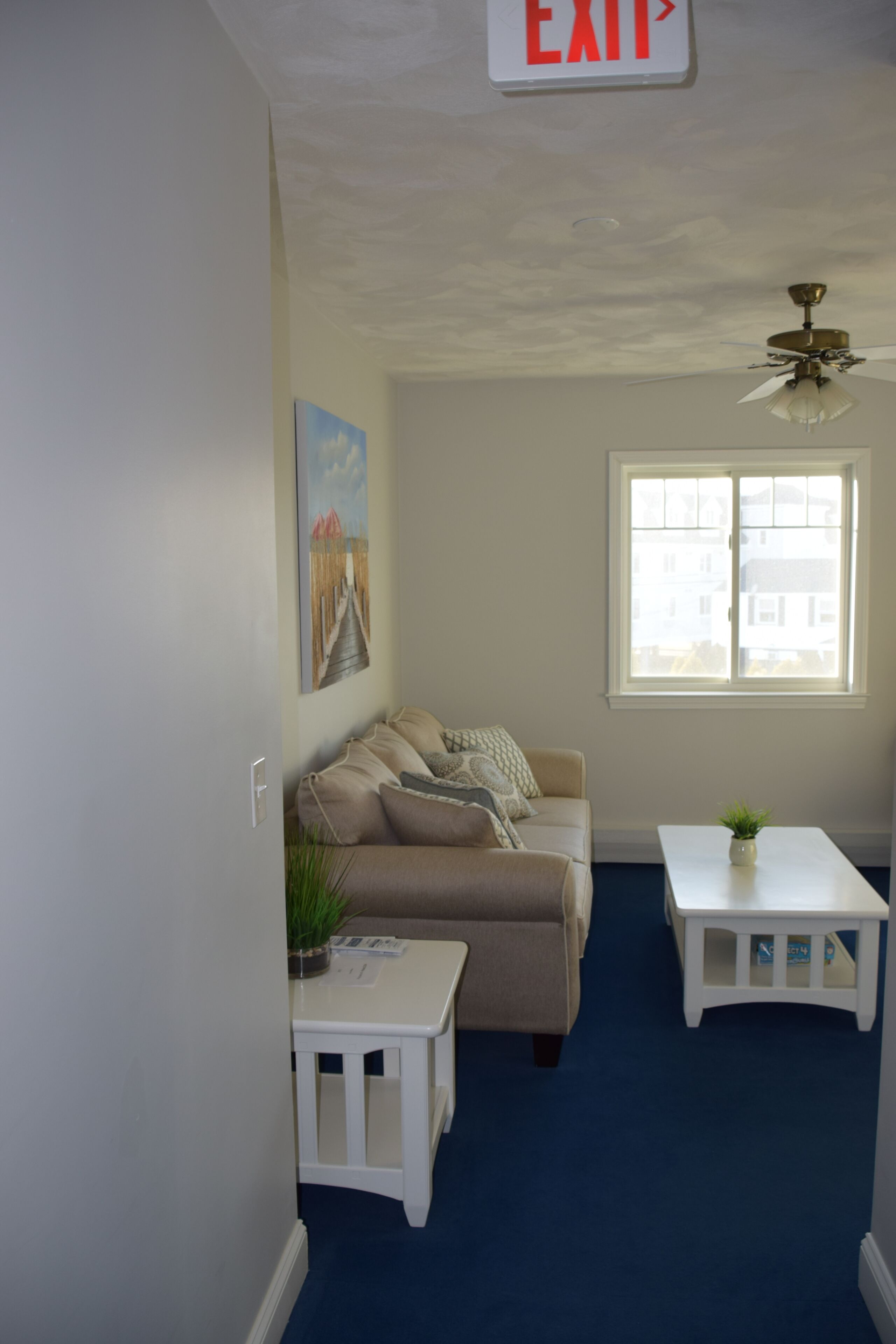 A living room with a beige sofa, a small white table, a ceiling fan, and a window with a view of buildings.