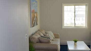 A living room with a beige sofa, a small white table, a ceiling fan, and a window with a view of buildings.