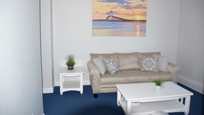 A living room with a beige sofa, white coffee table, and a painting of a sunset over water. - Rhea's Inn By The Sea (Middletown)