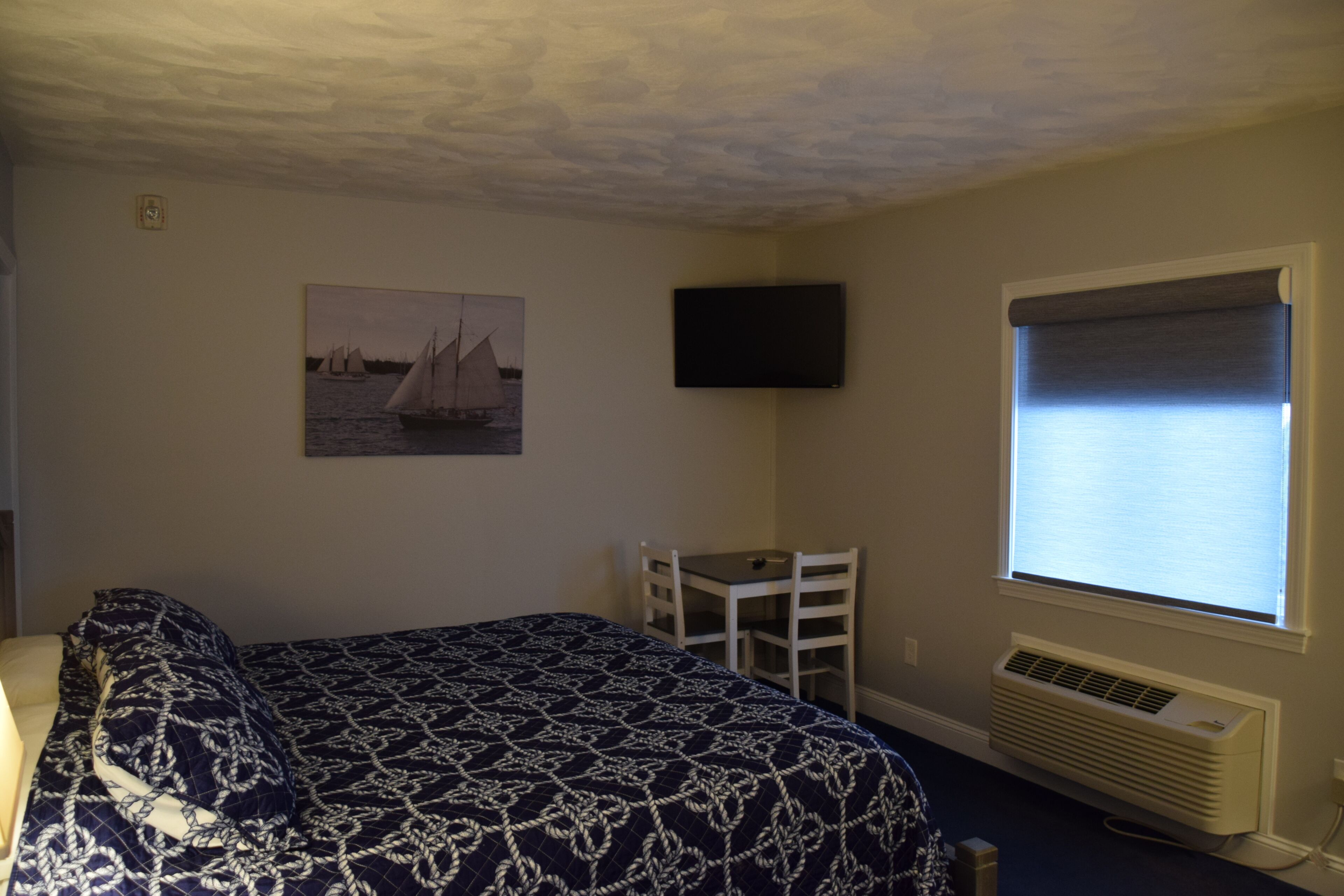 A bedroom with a bed, a desk, a chair, a television, and a window with blinds.