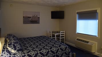 A bedroom with a bed, a desk, a chair, a television, and a window with blinds.