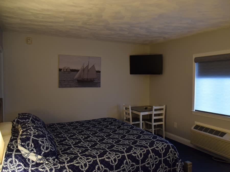 A bedroom with a bed, a desk, a chair, a television, and a window with blinds.