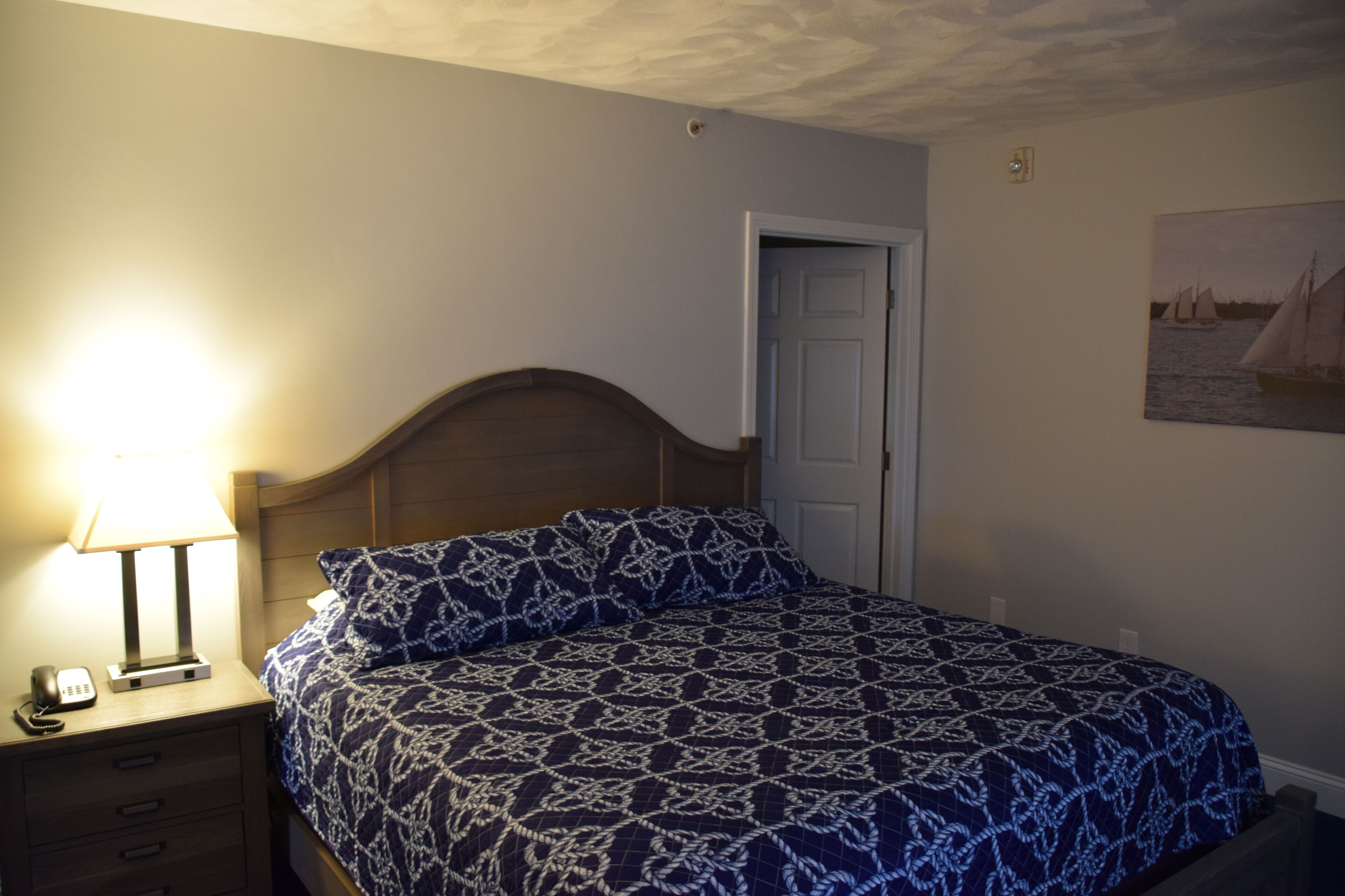 A bedroom with a bed, a nightstand with a lamp, a door, and a wall-mounted picture.