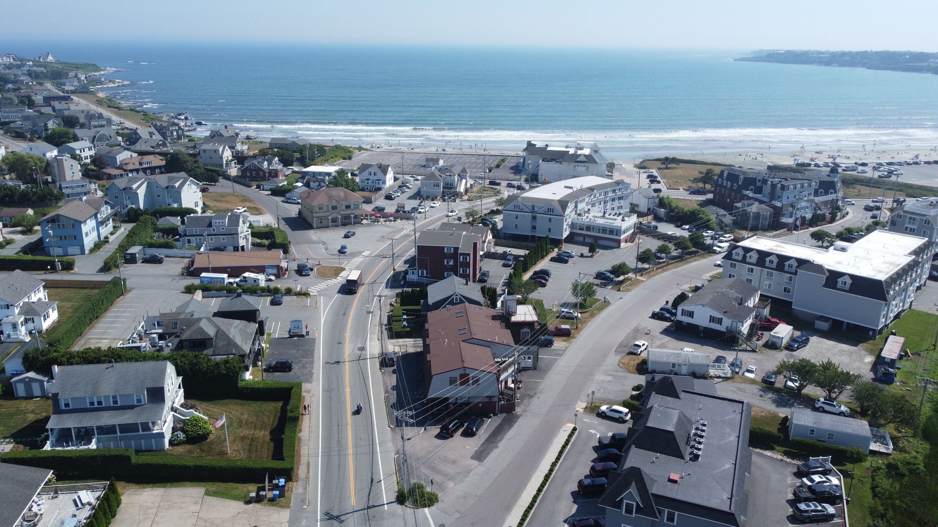 An aerial view of a coastal town with residential buildings, a main road, and a beach in the background.