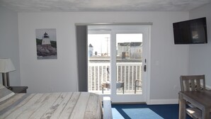 A room with a bed, a desk, a chair, a television, and a door leading to an outdoor area with a view of a building and a fence. - Rhea's Inn By The Sea (Middletown)
