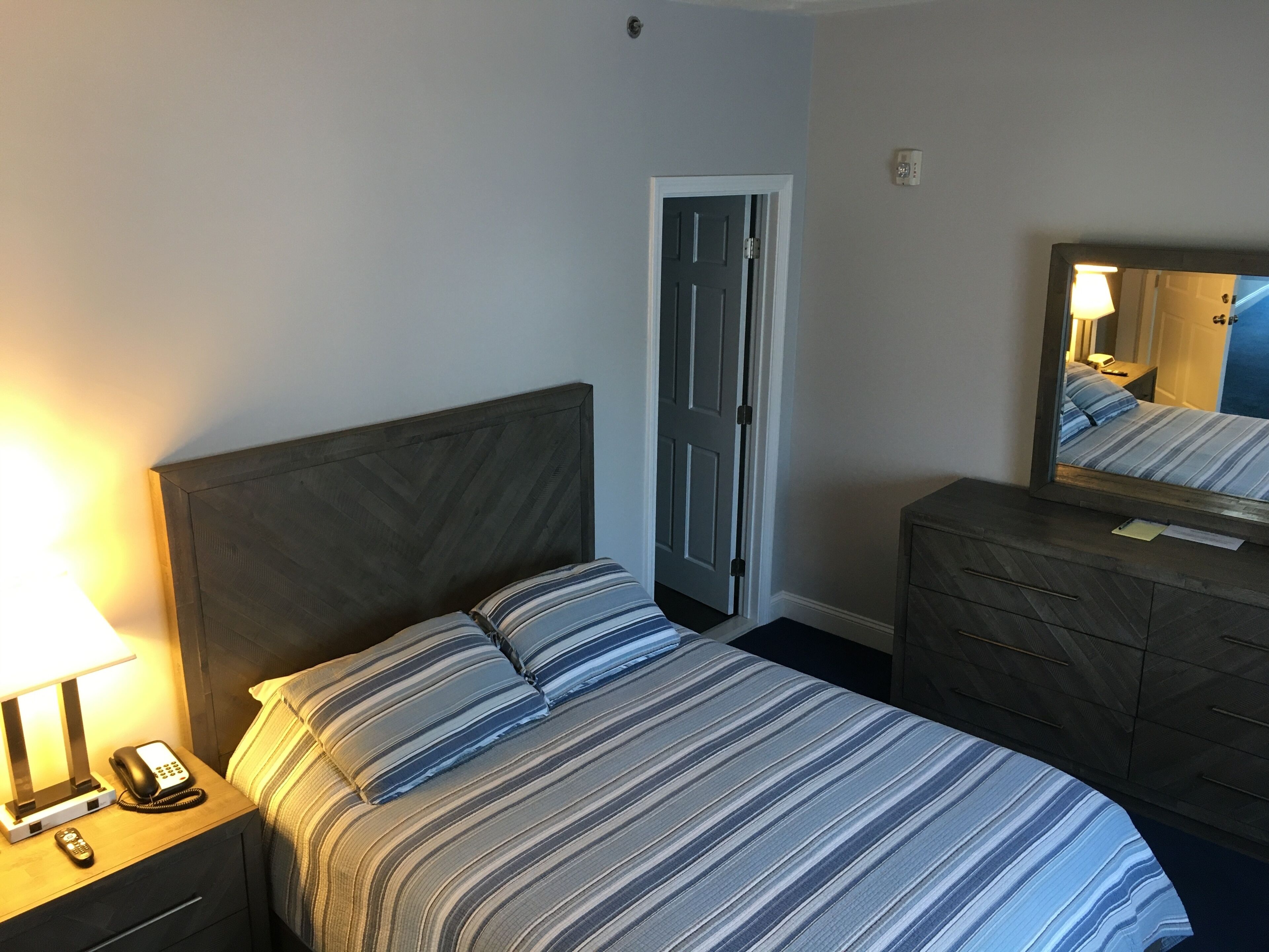 A hotel room with a bed, a nightstand with a lamp and phone, a dresser, and a door.