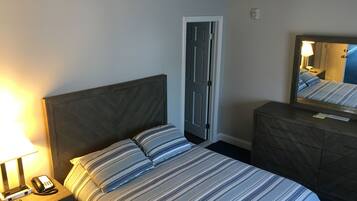 A hotel room with a bed, a nightstand with a lamp and phone, a dresser, and a door.