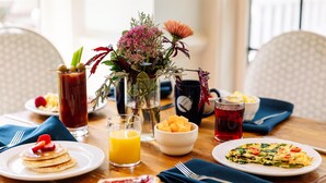 A breakfast table with pancakes, toast, a fruit bowl, a glass of orange juice, a pitcher of iced tea, and a vase of flowers.
