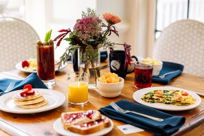 A breakfast table with pancakes, toast, a fruit bowl, a glass of orange juice, a pitcher of iced tea, and a vase of flowers.