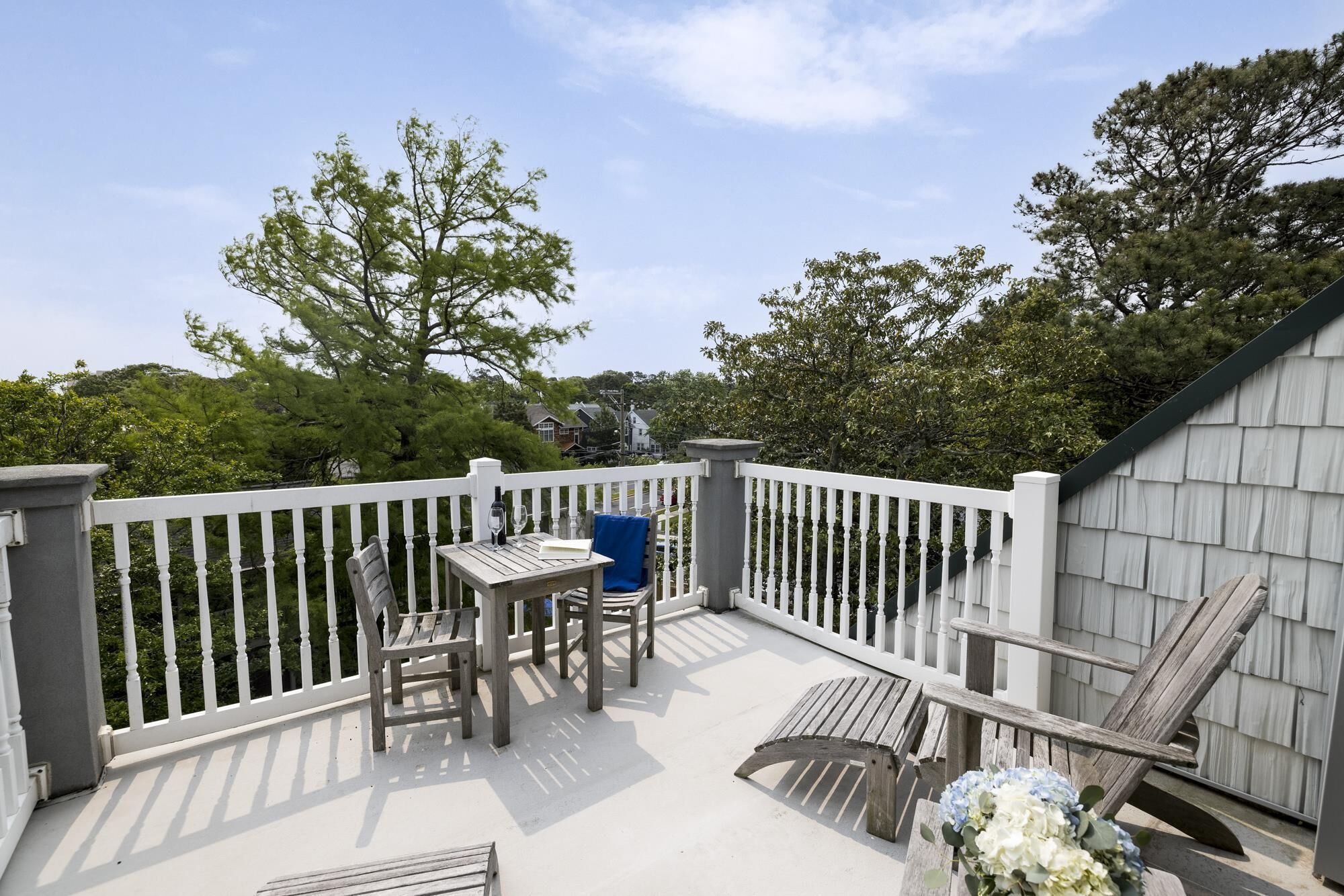 A covered outdoor deck with a table and chairs, surrounded by white railings and trees.