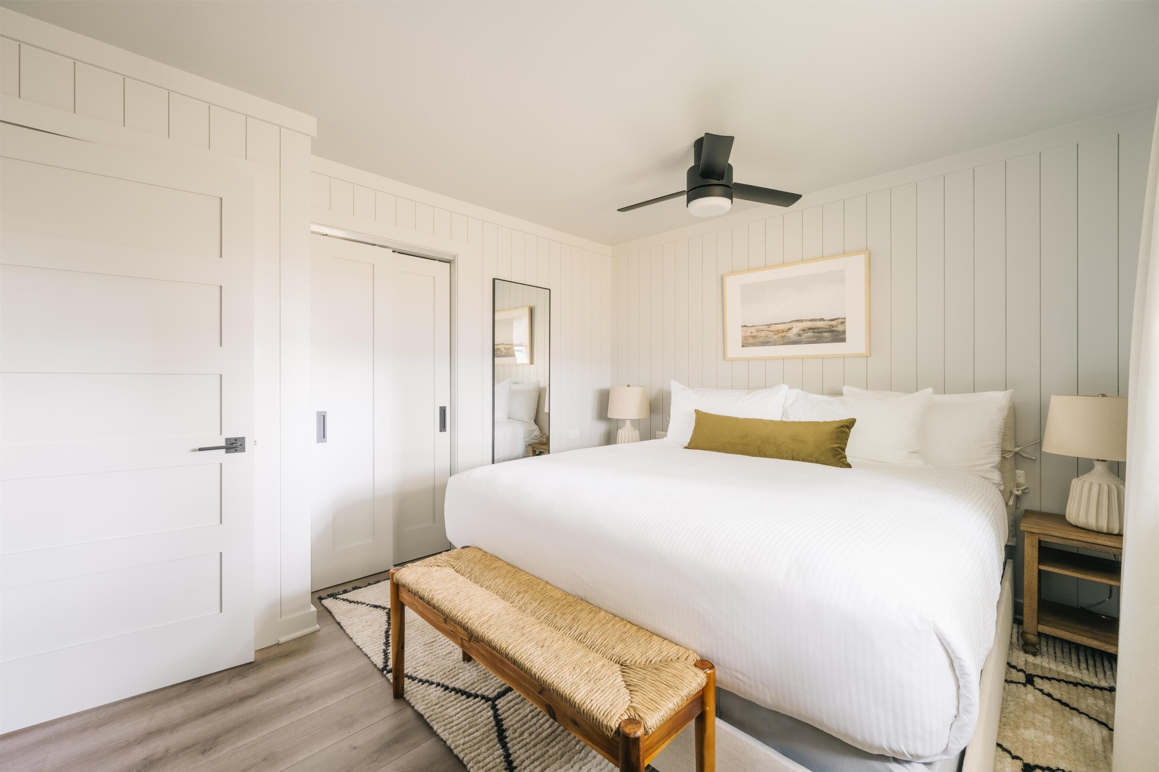 A bedroom with a large bed, a ceiling fan, a bench, and a framed picture on the wall.