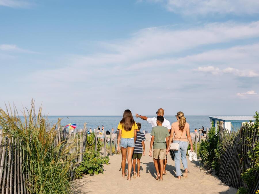 Een zandpad langs het strand met mensen die wandelen, een houten omheining en een heldere lucht.