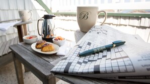 A wooden table with a crossword puzzle, a pen, a mug with a monogram, a coffee pot, and a plate of food.