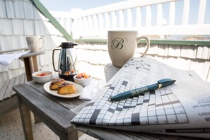 A wooden table with a crossword puzzle, a pen, a mug with a monogram, a coffee pot, and a plate of food.