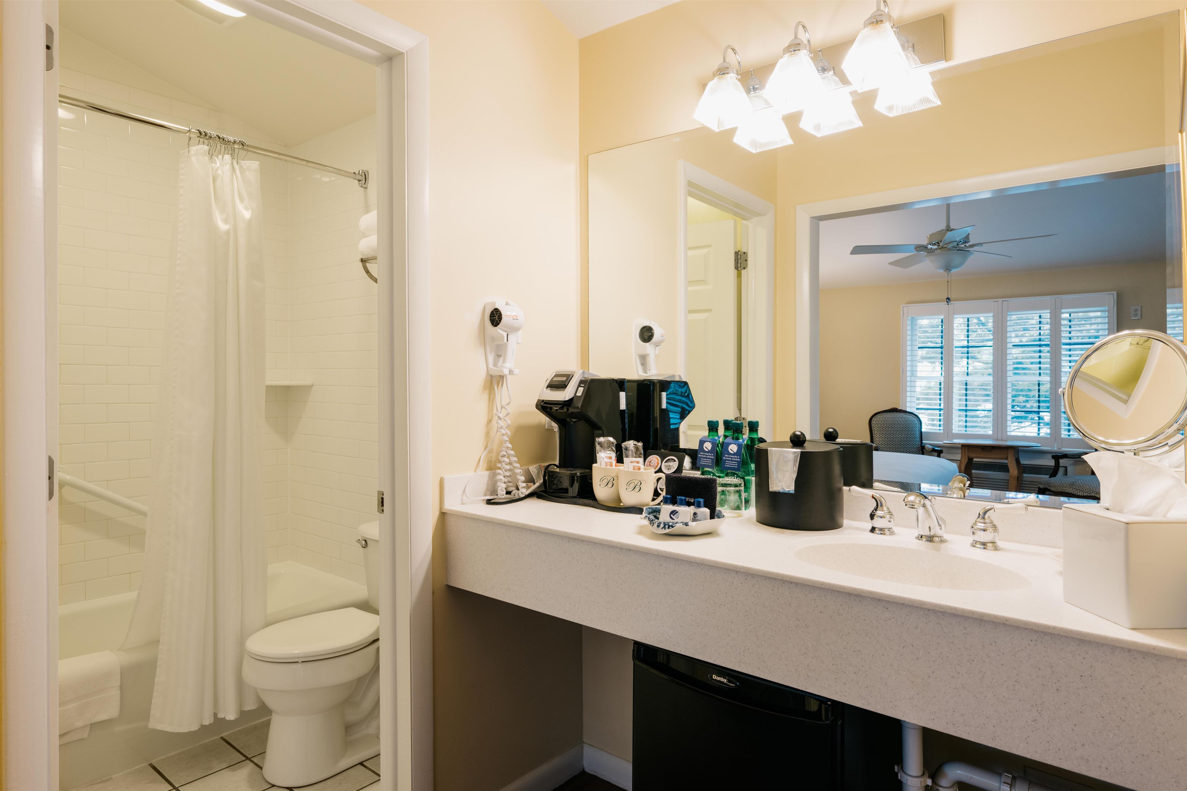 A bathroom with a shower, toilet, vanity with toiletries, and a mirror.
