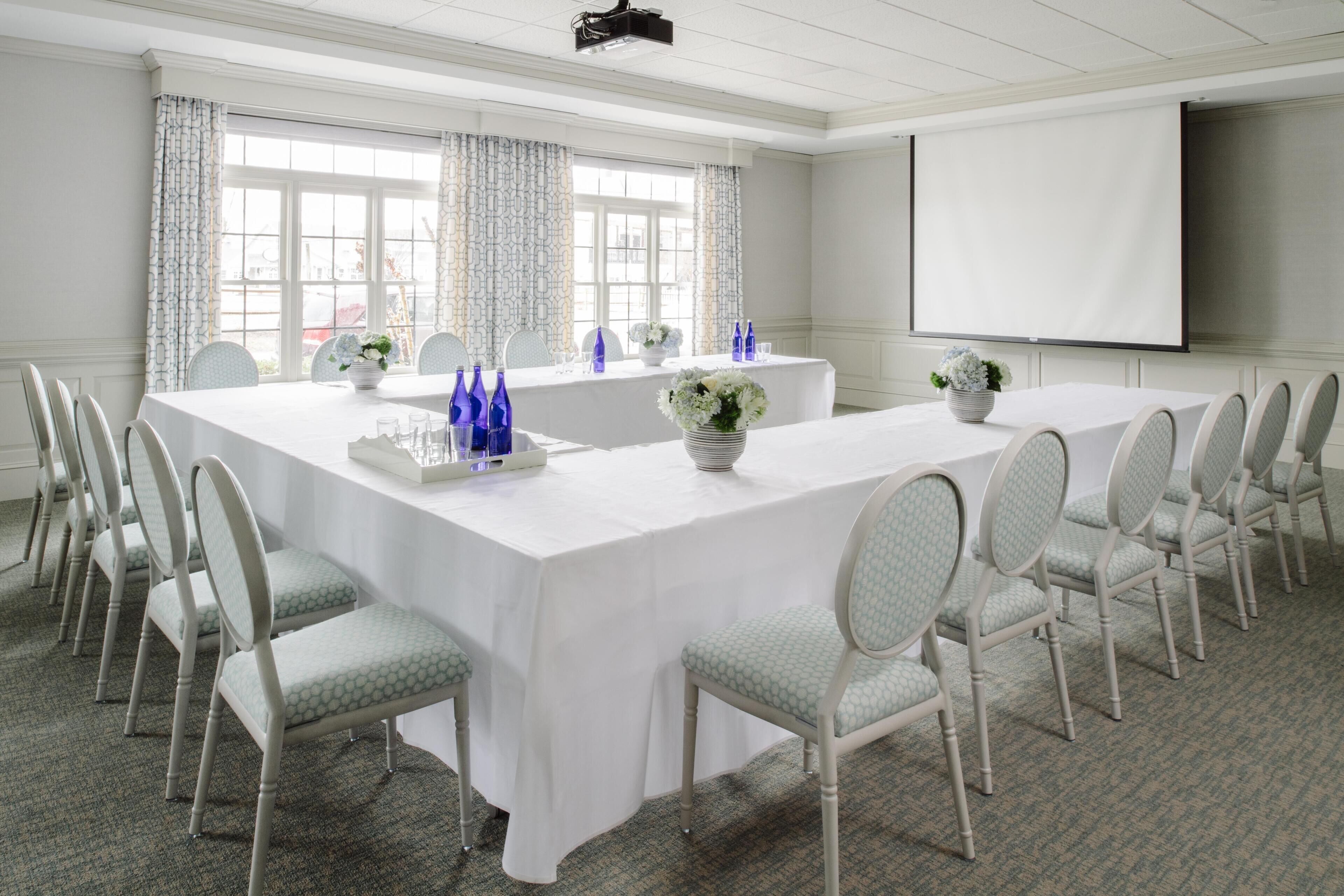 A conference room with a long table, white chairs, and a projector screen.